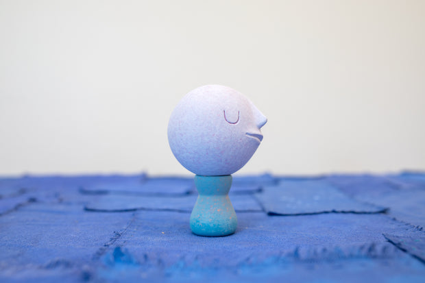 Small carved wooden sculpture of a purple moon with a closed eye, calm smiling expression. It rests atop of a blue wooden vessel shaped pedestal, like a kokeshi doll.