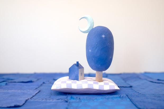 Diorama made up of small carved wooden sculptures depicting a small house next to a large oval shaped tree, with a crescent moon attached. They rest atop of a white and purple grid patterned wooden pillow, carved to look soft. Color scheme is purple, pink, blue and white.