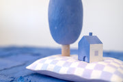 Diorama made up of small carved wooden sculptures depicting a small house next to a large oval shaped tree, with a crescent moon attached. They rest atop of a white and purple grid patterned wooden pillow, carved to look soft. Color scheme is purple, pink, blue and white.