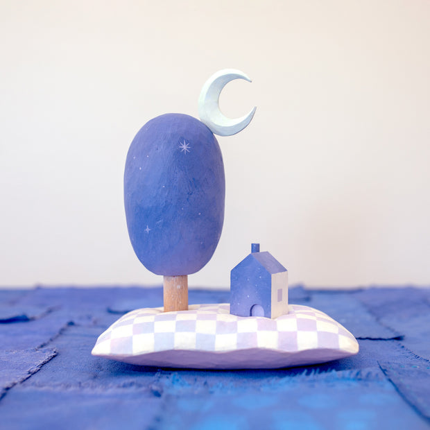 Diorama made up of small carved wooden sculptures depicting a small house next to a large oval shaped tree, with a crescent moon attached. They rest atop of a white and purple grid patterned wooden pillow, carved to look soft. Color scheme is purple, pink, blue and white.