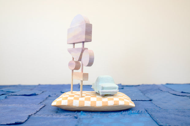 Diorama made up of small carved wooden sculptures depicting a light blue vintage car in front of several large sunset colored signs, attached to the same posts. They rest atop of a white and yellow grid patterned wooden pillow, carved to look soft. 