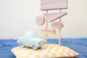 Diorama made up of small carved wooden sculptures depicting a light blue vintage car in front of several large sunset colored signs, attached to the same posts. They rest atop of a white and yellow grid patterned wooden pillow, carved to look soft. 