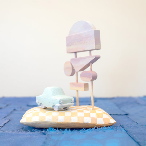 Diorama made up of small carved wooden sculptures depicting a light blue vintage car in front of several large sunset colored signs, attached to the same posts. They rest atop of a white and yellow grid patterned wooden pillow, carved to look soft. 