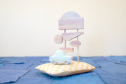 Diorama made up of small carved wooden sculptures depicting a light blue vintage car in front of several large sunset colored signs, attached to the same posts. They rest atop of a white and yellow grid patterned wooden pillow, carved to look soft. 