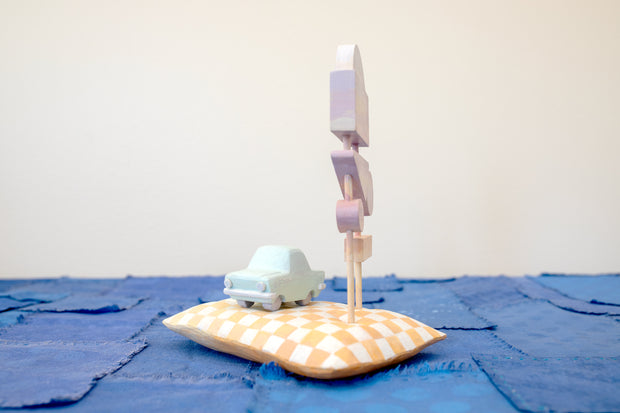 Diorama made up of small carved wooden sculptures depicting a light blue vintage car in front of several large sunset colored signs, attached to the same posts. They rest atop of a white and yellow grid patterned wooden pillow, carved to look soft. 