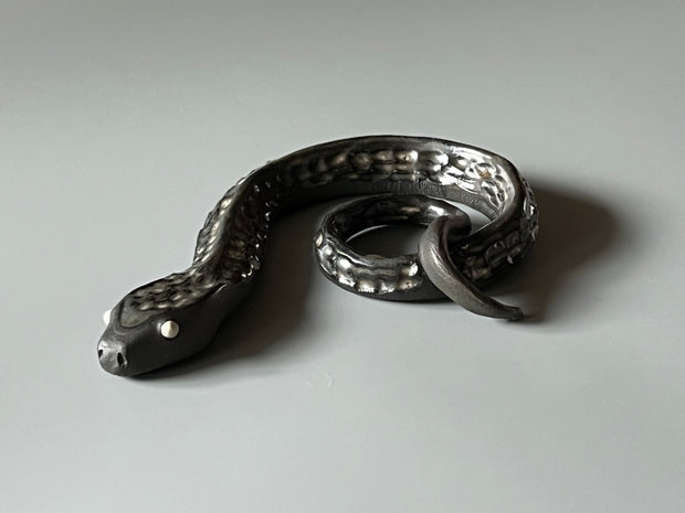 Ceramic sculpture of a dark silver snake with silver colored eyes and imprinted scales along its body. Its tail is curled into itself.