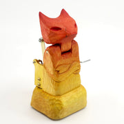 Whittled wooden sculpture of a cat or fox-like devil with its arms crossed. It is painted in a red to yellow gradient and has a mechanical turning element to it.