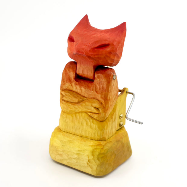 Whittled wooden sculpture of a cat or fox-like devil with its arms crossed. It is painted in a red to yellow gradient and has a mechanical turning element to it.