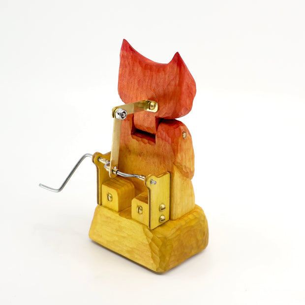 Whittled wooden sculpture of a cat or fox-like devil with its arms crossed. It is painted in a red to yellow gradient and has a mechanical turning element to it.