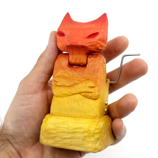 Whittled wooden sculpture of a cat or fox-like devil with its arms crossed. It is painted in a red to yellow gradient and has a mechanical turning element to it.