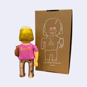 Back view of vinyl figure of a man with a receding hairline but long gold hair. He holds a bonito fish and wears a pink shirt that says "SHIT!" on the back. He stands next to a cardboard box with a line art illustration of himself on it.