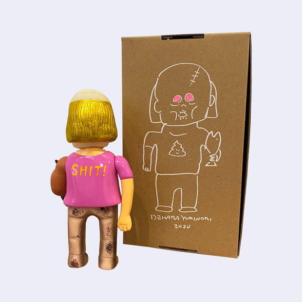 Back view of vinyl figure of a man with a receding hairline but long gold hair. He holds a bonito fish and wears a pink shirt that says "SHIT!" on the back. He stands next to a cardboard box with a line art illustration of himself on it.