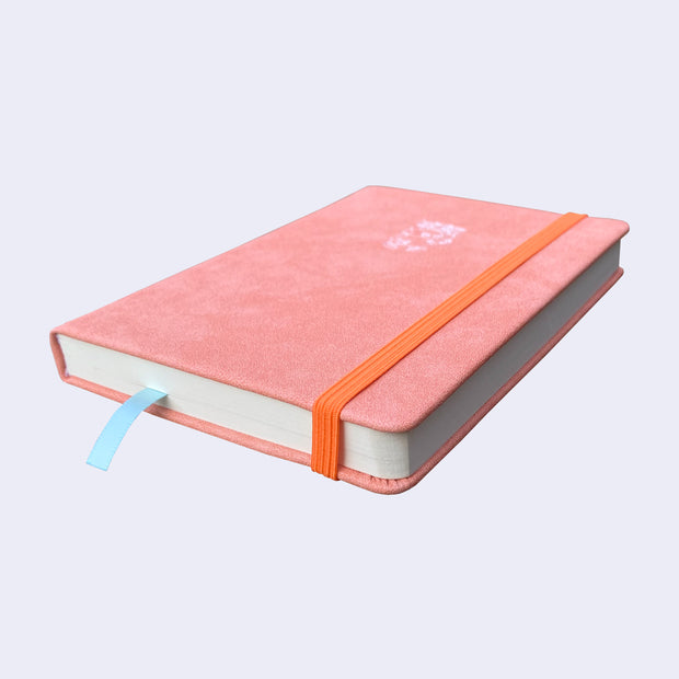 Peach colored pleather covered sketchbook with a small embossment of a robot carrying a flag that reads "Giant Robot" in the upper center. A bright orange elastic band keeps the sketchbook closed. Side view.