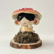 Ceramic sculpture of a small character with dark sunglasses standing on a pile of moss atop a wooden bark circle. Atop their head is a red mushroom cap with white dots.