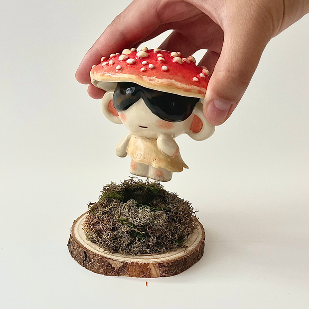 Ceramic sculpture of a small character with dark sunglasses standing on a pile of moss atop a wooden bark circle. Atop their head is a red mushroom cap with white dots.