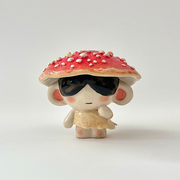 Ceramic sculpture of a small character with dark sunglasses standing on a pile of moss atop a wooden bark circle. Atop their head is a red mushroom cap with white dots.