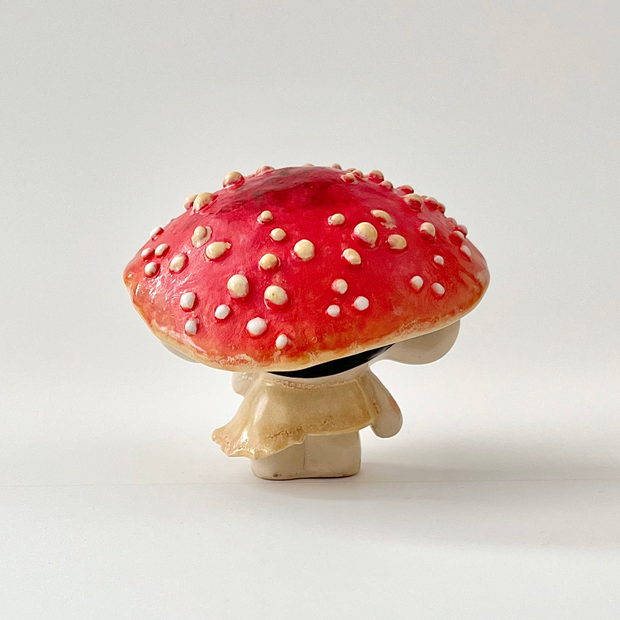 Ceramic sculpture of a small character with dark sunglasses standing on a pile of moss atop a wooden bark circle. Atop their head is a red mushroom cap with white dots.