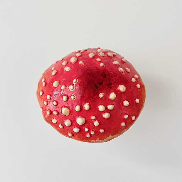 Ceramic sculpture of a small character with dark sunglasses standing on a pile of moss atop a wooden bark circle. Atop their head is a red mushroom cap with white dots.