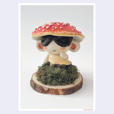 Photograph of a ceramic sculpture of a small character with dark sunglasses standing on a pile of moss atop a wooden bark circle. Atop their head is a red mushroom cap with white dots.