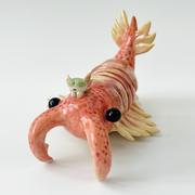 Ceramic sculpture of a large crustacean with 2 front prongs, a feathered tail and feathered gils. It has large black eyes at each side and a small character sits atop its head.