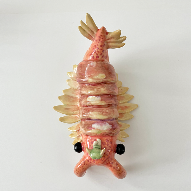 Ceramic sculpture of a large crustacean with 2 front prongs, a feathered tail and feathered gils. It has large black eyes at each side and a small character sits atop its head.