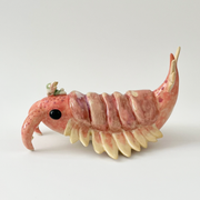 Ceramic sculpture of a large crustacean with 2 front prongs, a feathered tail and feathered gils. It has large black eyes at each side and a small character sits atop its head.