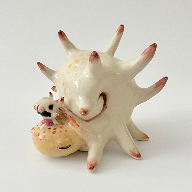 Ceramic sculpture of a cream colored shelled bug or crustacean, akin to a snail with a spiked and rounded shell. A small character sits atop its head.
