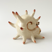 Ceramic sculpture of a cream colored shelled bug or crustacean, akin to a snail with a spiked and rounded shell. A small character sits atop its head.