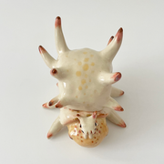 Ceramic sculpture of a cream colored shelled bug or crustacean, akin to a snail with a spiked and rounded shell. A small character sits atop its head.