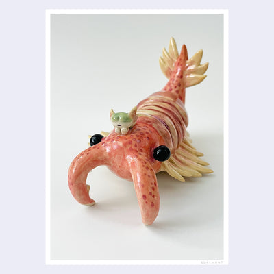 Photograph of a ceramic sculpture of a pinkish red crustacean, akin to a shrimp or krill. It has a small character sitting atop its head.