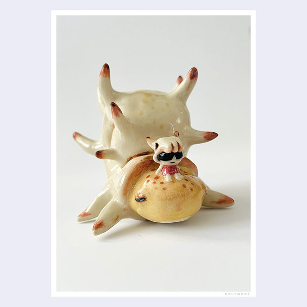 Photograph of a ceramic sculpture of a snail like crustacean, with a white spiked shell and a small character sitting atop its head.