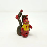 Small ceramic sculpture of a red devil character with sharp yellow teeth, yellow eyes and a yellow t-shirt with a red sun on it. It stands in front of a set of nopales with red blooms.