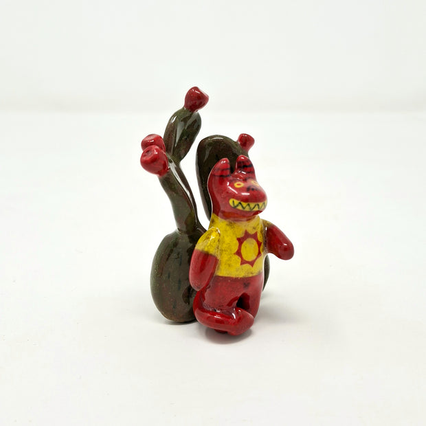 Small ceramic sculpture of a red devil character with sharp yellow teeth, yellow eyes and a yellow t-shirt with a red sun on it. It stands in front of a set of nopales with red blooms.