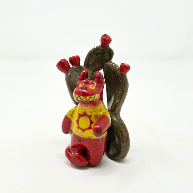 Small ceramic sculpture of a red devil character with sharp yellow teeth, yellow eyes and a yellow t-shirt with a red sun on it. It stands in front of a set of nopales with red blooms.