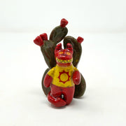 Small ceramic sculpture of a red devil character with sharp yellow teeth, yellow eyes and a yellow t-shirt with a red sun on it. It stands in front of a set of nopales with red blooms.