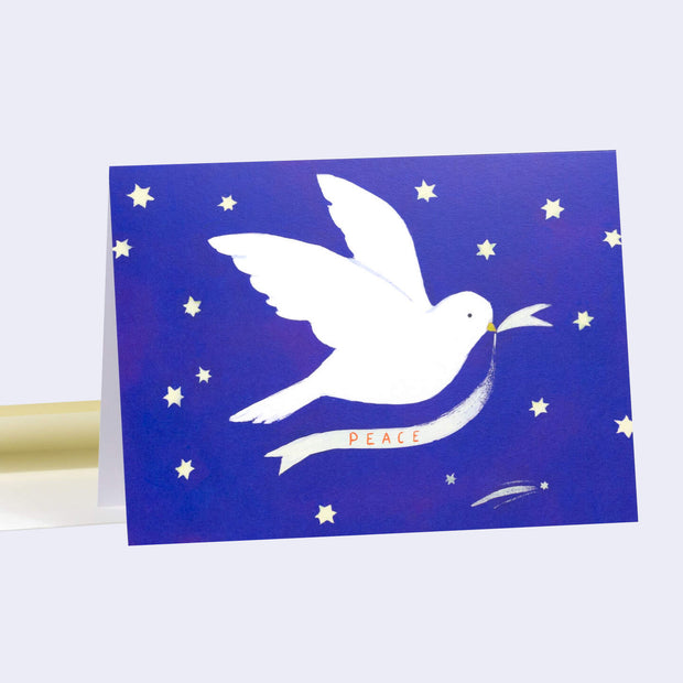 Holiday card with blue background and an illustration of a white dove flying and holding a small ribbon of fabric that reads "peace".