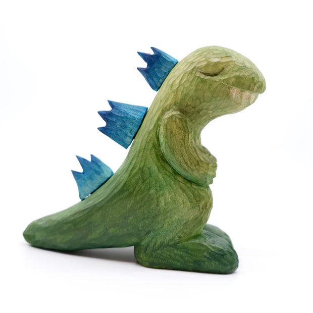 Whittled wooden sculpture of a green Godzilla with blue back spikes, modeled in a non aggressive, cute fashion. Its eyes are peacefully closed and its hands rest on its chest.