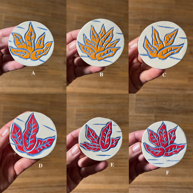 Collage of 6 ceramic circular brooches. Each feature a simple carved illustration of a sea plant, 3 are colored light orange and 3 are colored red.