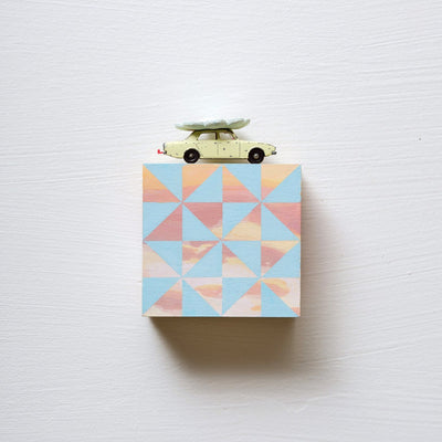 Small square wood panel painted like an orange-pink sunset with blue patterning overlayed on top, akin to a quilt tile design. Atop of it is a painted toy car with a wooden flower on top of its roof.