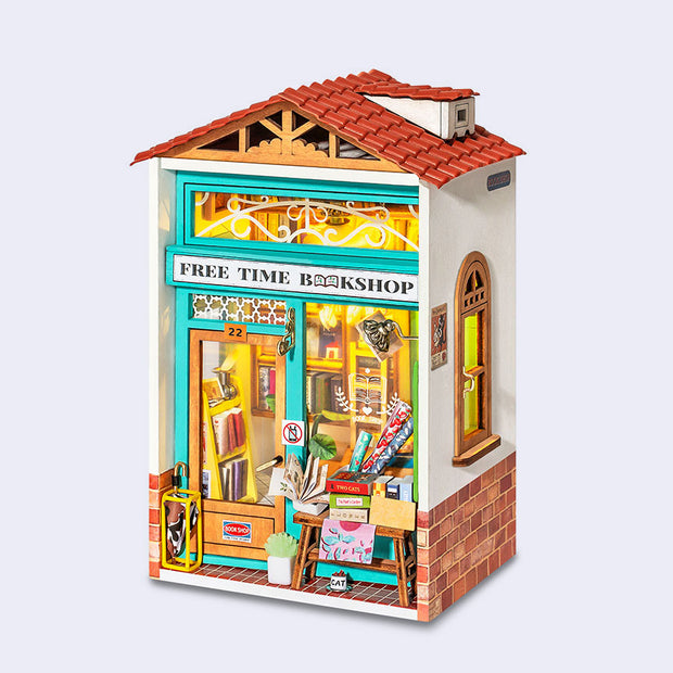 Assembled diorama of a miniature bookshop, with lights on inside and books visible from the windows. Outside are various sale items and wrapping paper.