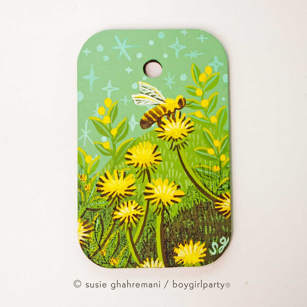 Painting on rounded corner wood panel of a bumble bee on top of a yellow dandelion. Background is mint green with more grass and yellow flowers.