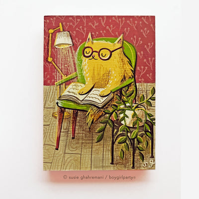 Painting on wood of a fluffy yellow cartoon cat sitting in a green armchair and reading a book, with a lamp shining down onto it. A houseplant is on a side table nearby.