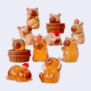 10 small plastic figures of orange or brown capybaras, all with their eyes closed and a calm expression. They are in varying poses of relaxation and some have citrus atop of their head.