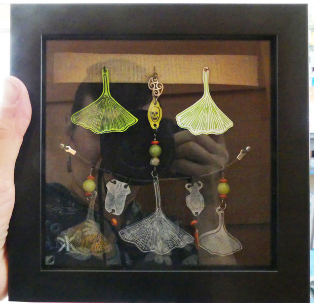 Collage on black fabric of colored plastic pieces, mostly upside down gingko leaves with beads and charms.