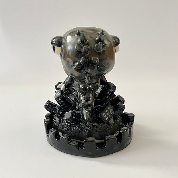 Ceramic sculpture, mostly a glossy black with a multicolored sheen of a character dressed in a Gojira costume, with claws and only the character's face peeping out. It stands atop a large pile of buildings.