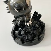 Ceramic sculpture, mostly a glossy black with a multicolored sheen of a character dressed in a Gojira costume, with claws and only the character's face peeping out. It stands atop a large pile of buildings.