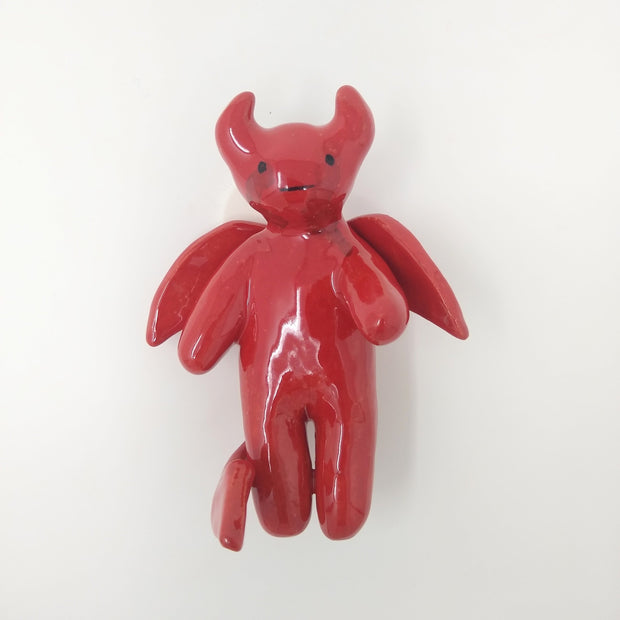 Ceramic sculpture of a red cute cartoon devil, with mostly sooth body features and small horns and wings.