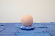 Small carved wooden sculpture of a tannish orange sphere with a closed eye, calm smiling expression. It rests atop of a rounded purple wooden pillow with splatter patterns, carved to look soft.