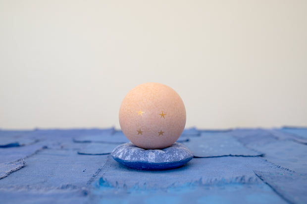 Small carved wooden sculpture of a tannish orange sphere with a closed eye, calm smiling expression. It rests atop of a rounded purple wooden pillow with splatter patterns, carved to look soft.