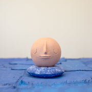 Small carved wooden sculpture of a tannish orange sphere with a closed eye, calm smiling expression. It rests atop of a rounded purple wooden pillow with splatter patterns, carved to look soft.